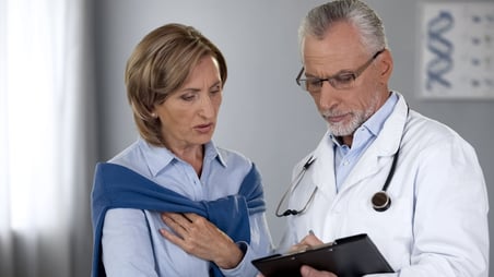 A doctor wearing a stethoscope and lab coat takes notes as a female patient stands by and watches