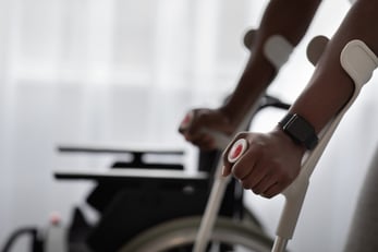 A person’s arms grip the handles of a walker in front of a blurred image of a wheelchair