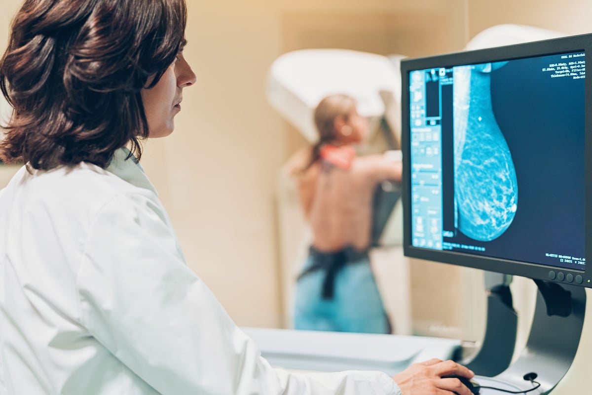 A physician views an image on a computer screen, while a woman in the background has a mammogram