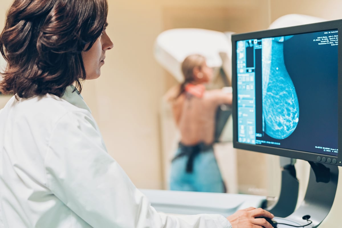 Un médico observa una imagen en la pantalla de un ordenador, mientras una mujer al fondo se hace una mamografía.