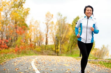 A smiling woman jogs down a road surrounded by fall foliage