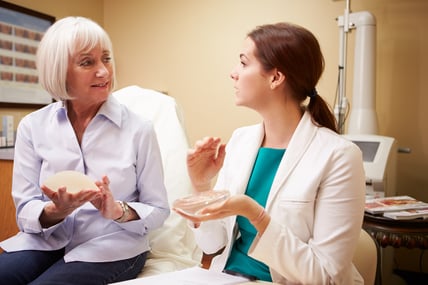 A young clinician and older woman holding different types of breast implants are sitting inside a medical facility, having a discussion