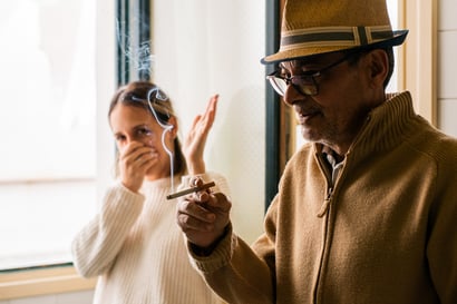 Elderly man smoking a cigarette oblivious to the discomfort the smoke produces on a young woman