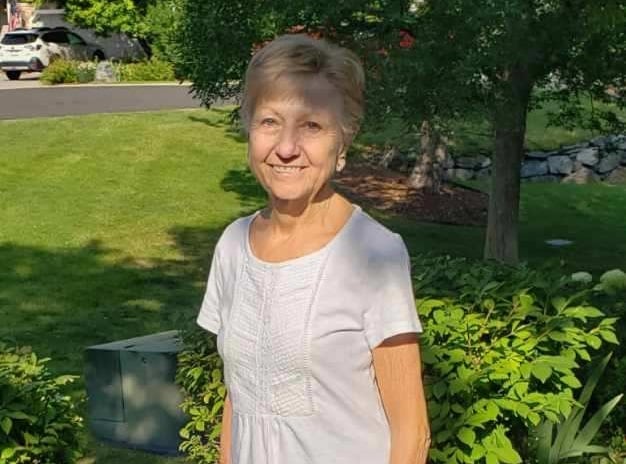 A woman in a white top stands outside in her yard and smiles at the camera.