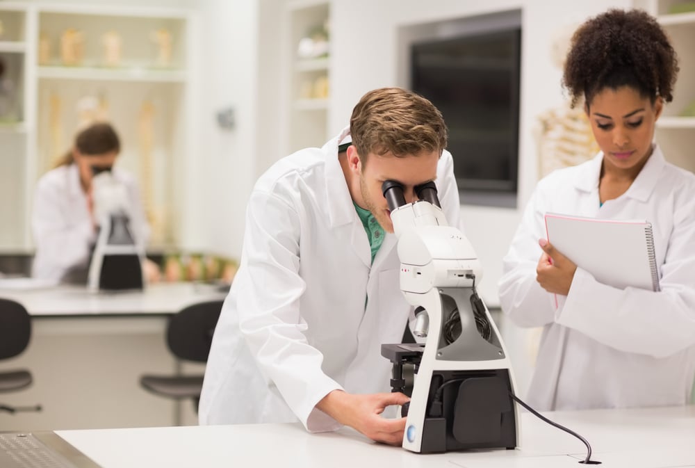 Medical students are in lab. One student looks in a microscope while the other one takes notes.