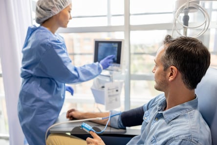 A male patient sits in a chair while having his vital signs checked. A health care provider wearing a blue gown and hair net analyzes data on a computer screen.