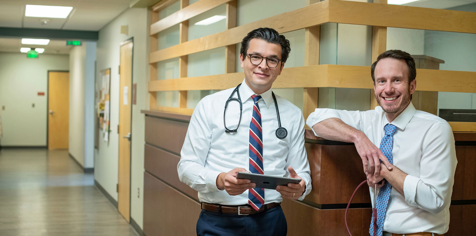Two doctors pose in a hospital hallway and smile at the camera.