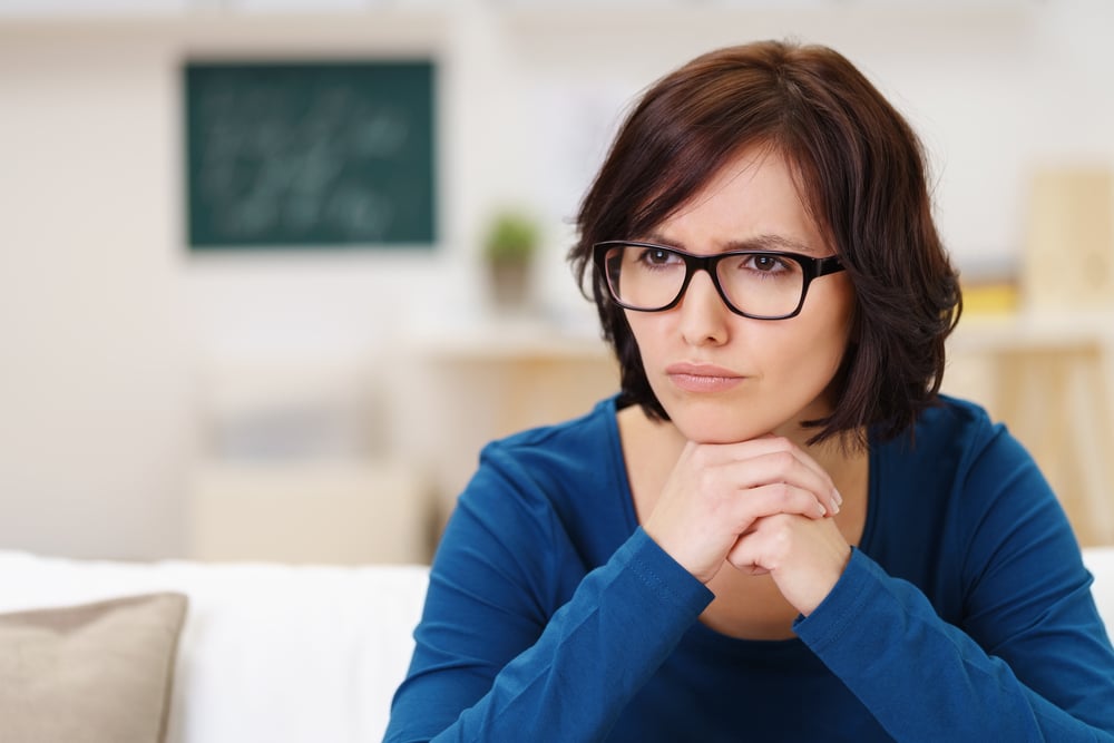 Mujer pensativa con gafas, sentada en el sofá del salón y mirando hacia otro lado con la barbilla apoyada en las manos.