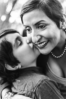 A smiling, middle-aged woman with short hair and a pearl necklace bends down to receive a kiss on the cheek from a child
