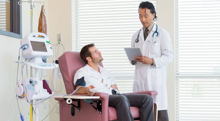 A doctor in a lab coat holds a clipboard and speaks to a man sitting in a chair receiving chemotherapy