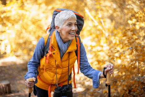 A woman wearing a backpack leans forward in front of autumn trees