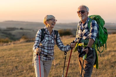 a_mature_couple_wearing_backpacks_and_holding_walking_sticks_are_on_top_of_a_mountain_and_laughing