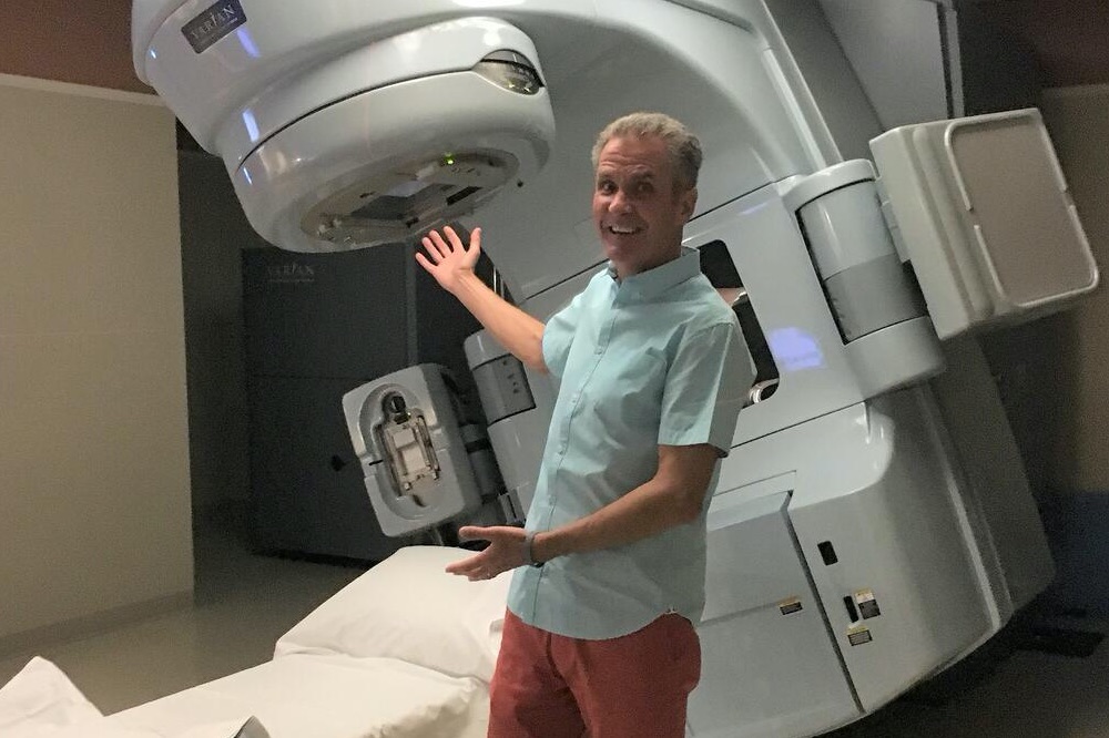 A man stands in front of a radiation machine and smiles