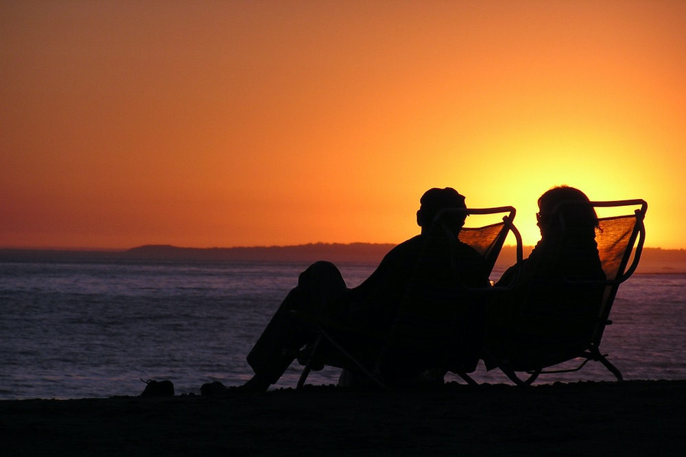 Two people sit on the beach in low beach chairs in front of a setting sun.