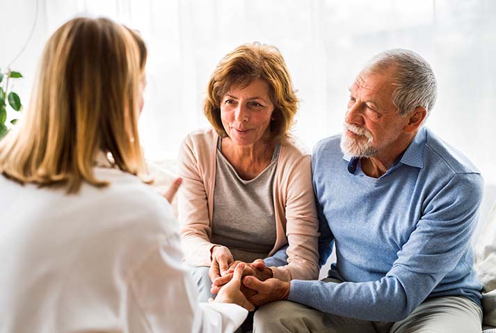 A man and woman sit next to each other across from a medical provider who is speaking to them. The man and woman are holding hands and the medical provider is reaching out to their hands.