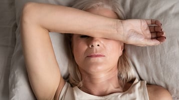 Fatigued woman lays in bed with arms over face